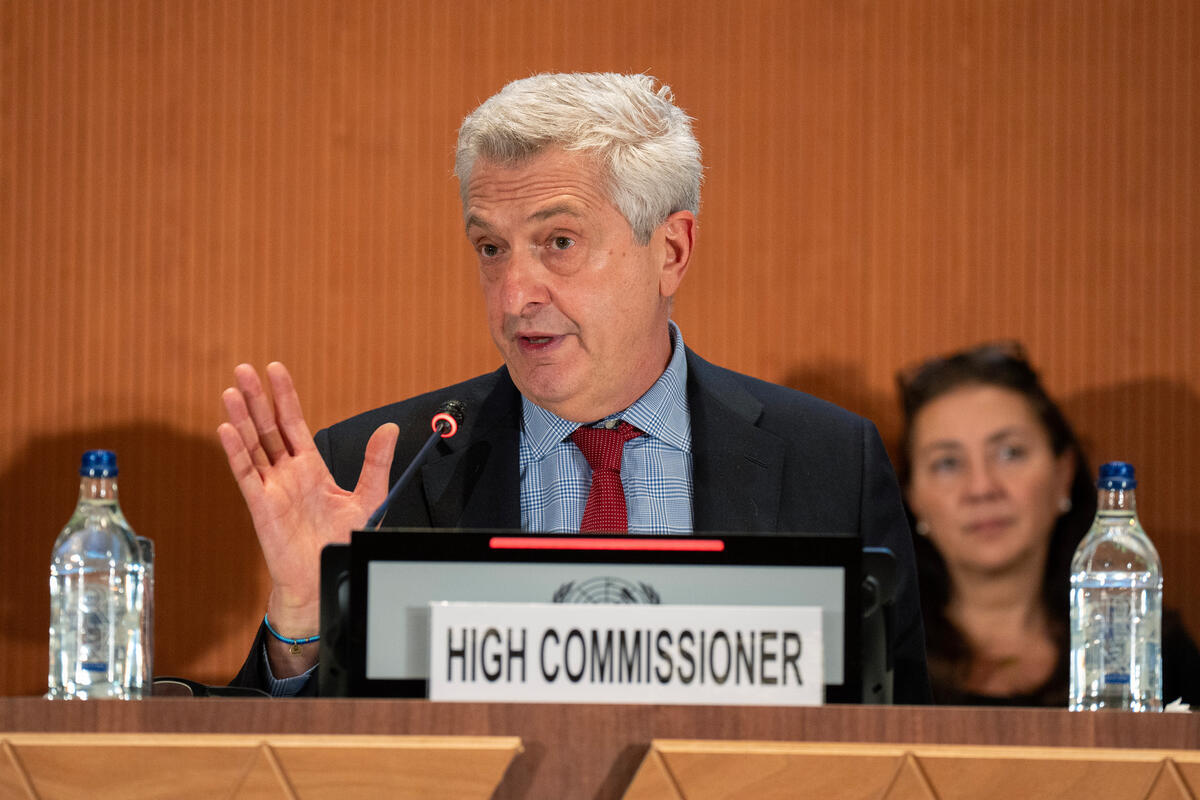 A man sits behind a dais making a speech with a sign reading 'High Commissioner' in front of him