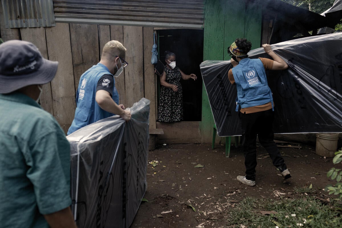 UNHCR staff deliver mattresses to a woman who stands in a doorway of a shelter.