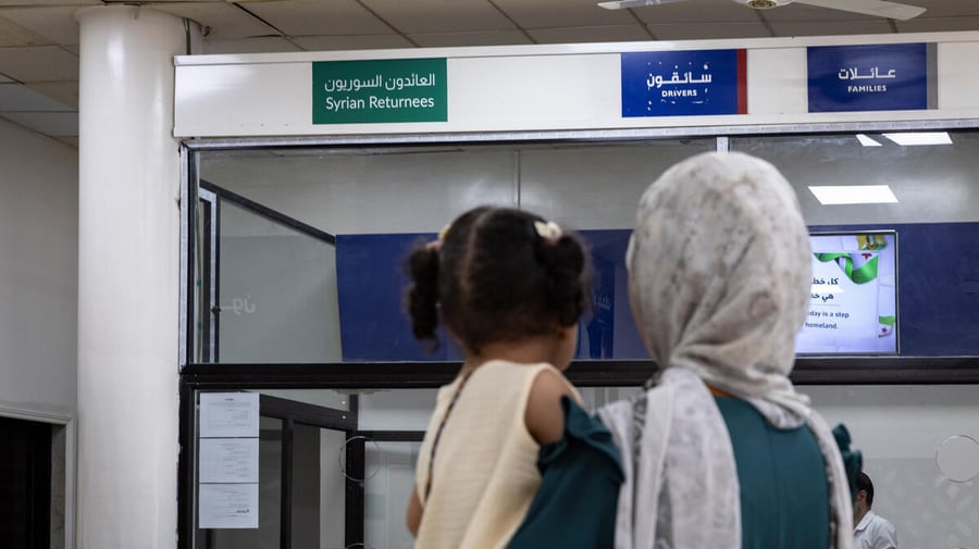 A woman carrying her daughter stands in front of a reception counter with a sign reading 'Syrian returnees' in Arabic and English