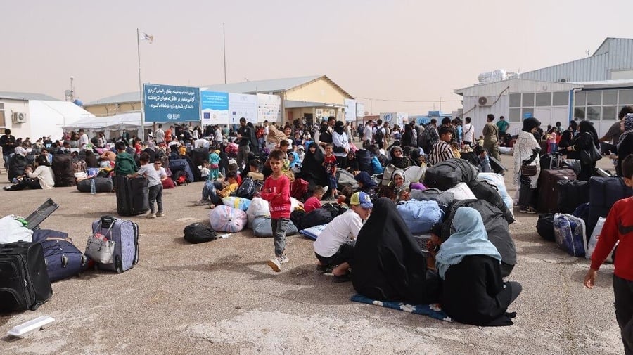 Families with their belongings gather at a border crossing, standing or sitting on the ground.
