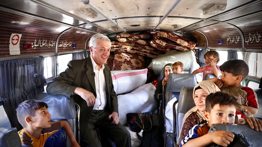 A smiling man in a suit stands inside a bus with women and children seated around him and mattresses piled in the rear of the vehicle High Commissioner for Refugees Filippo Grandi visits the Jdeidat/Yabous border to mark World Refugee Day 2025.