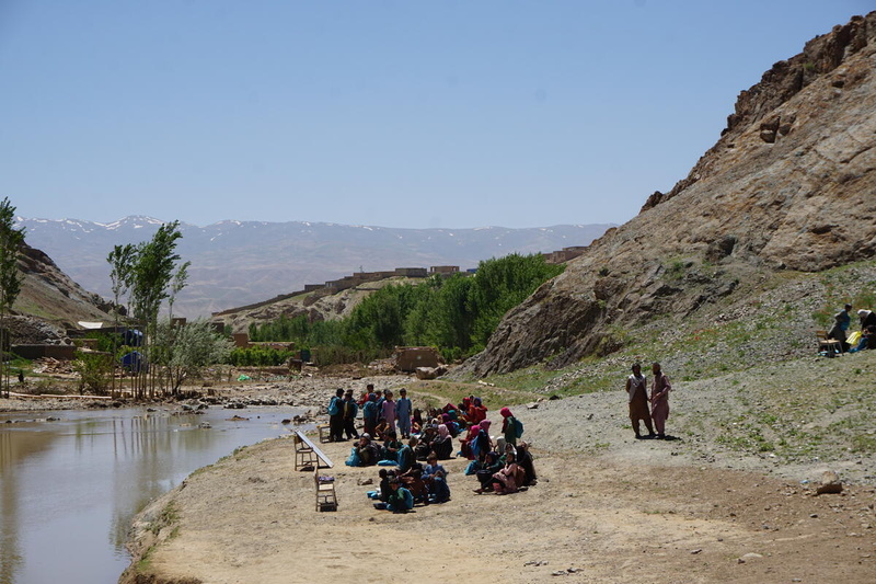 Children sit ouside on a patch of earth at the bottom of a hillside next to a pool of floodwater