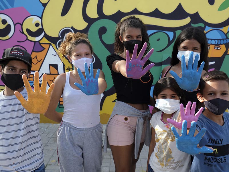 A group of six children and youth standing in front of a mural, showing their hands covered I paint, and smiling with their masks on 