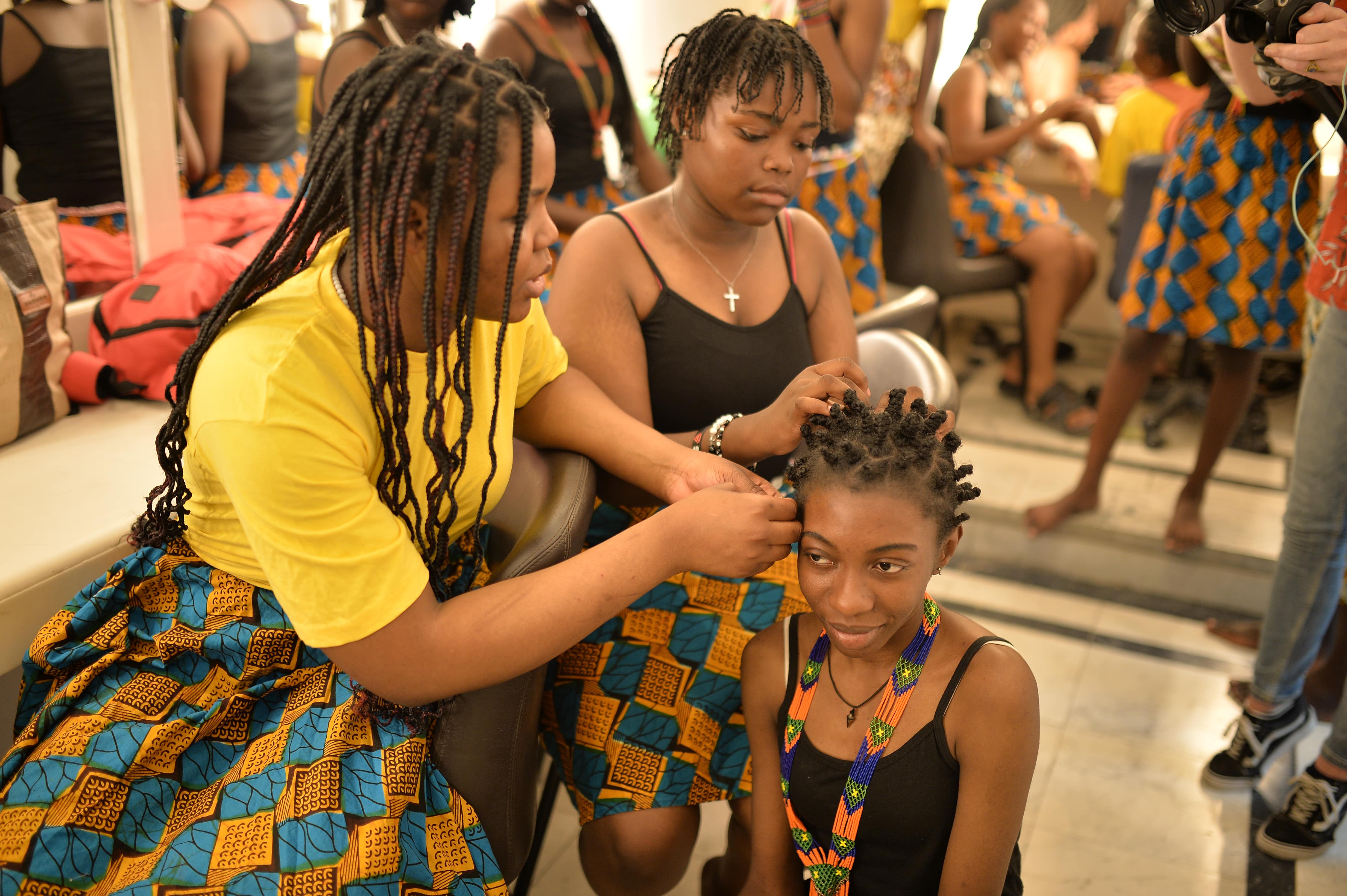 Two young women braid another's hair backstage.