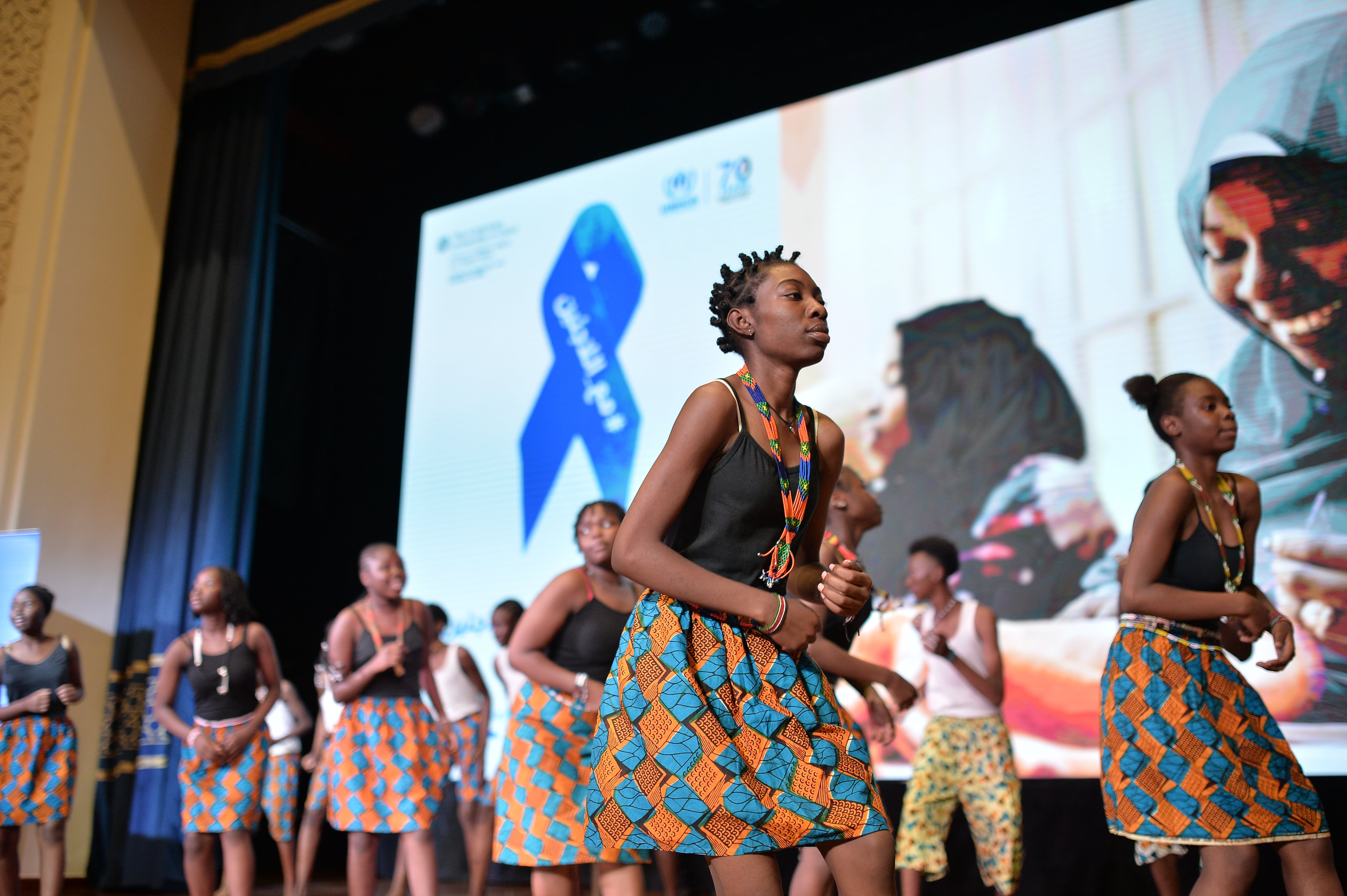 Dancers perform on stage in front of a large projection of a World Refugee Day ribbon.