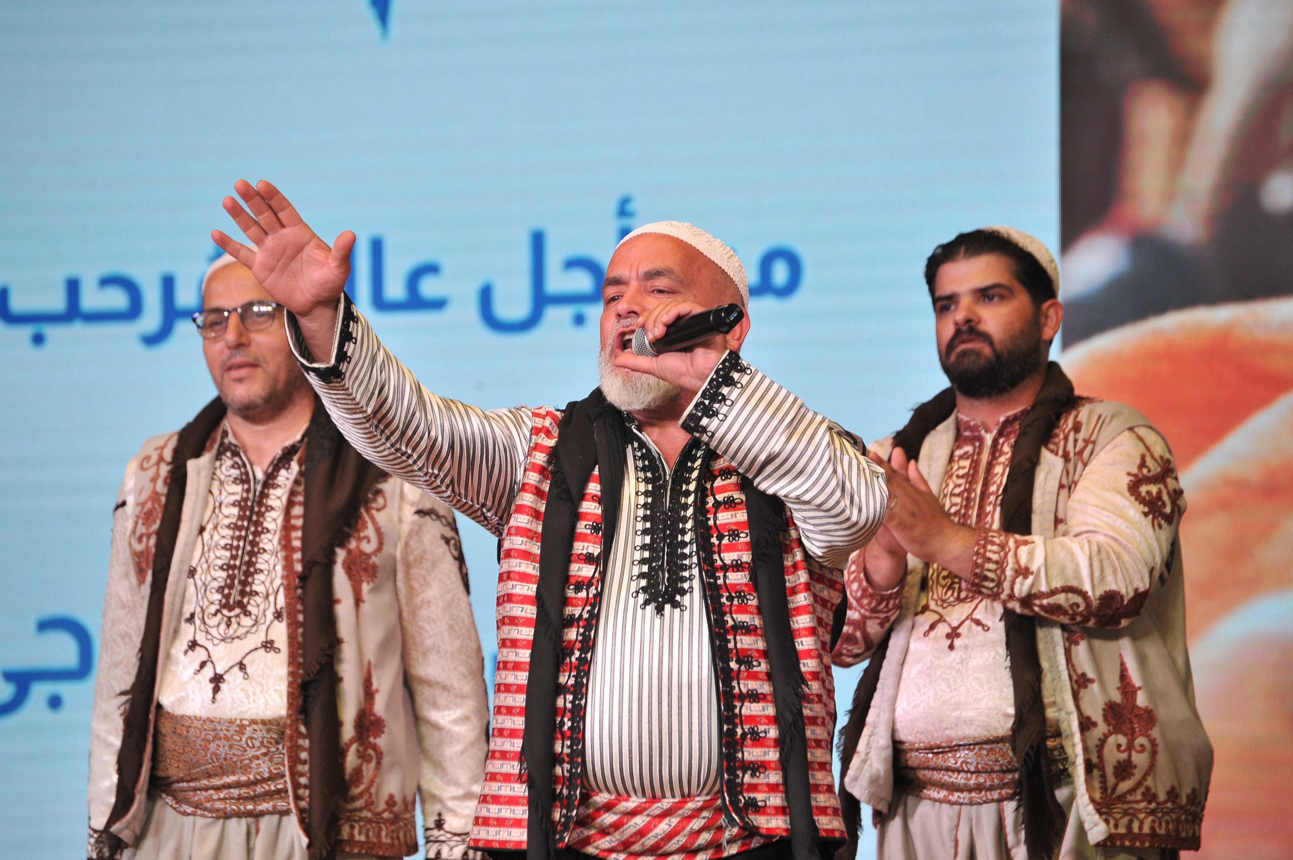 A man wearing traditional costume sings into a microphone and raises a hand to the audience while two other performers stand either side of him