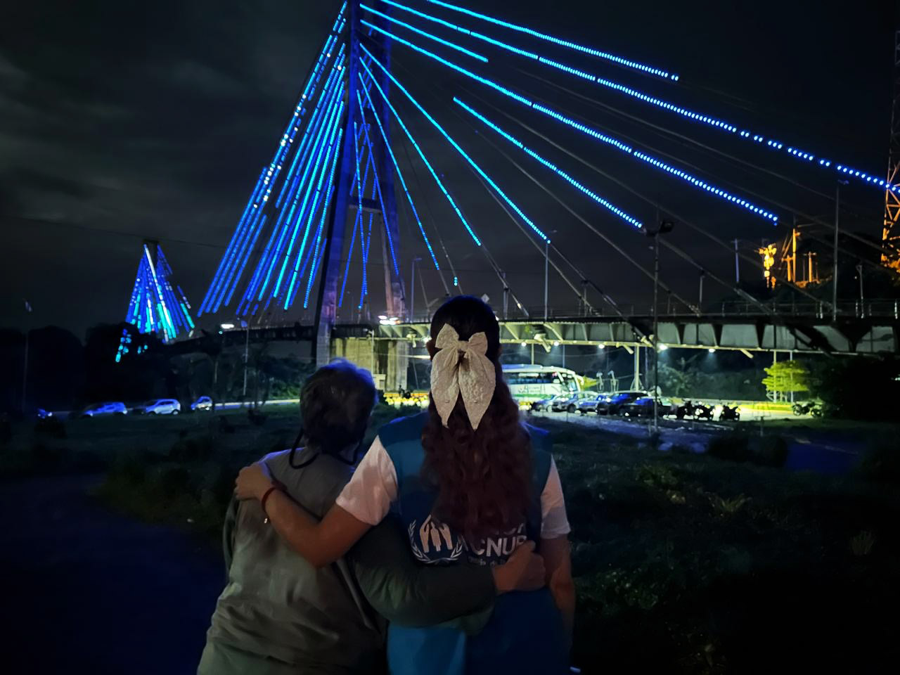 Two women embrace in front of a bridge lit in blue at night