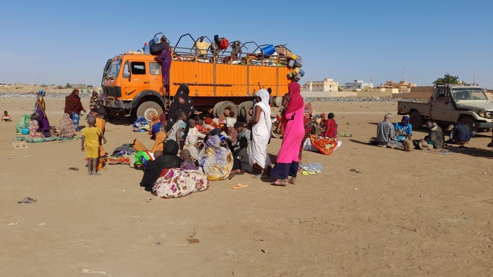 Families of displaced people gather with their belongings in front of and on top of a large truck, in an arid environment, with a utility vehicle nearby.