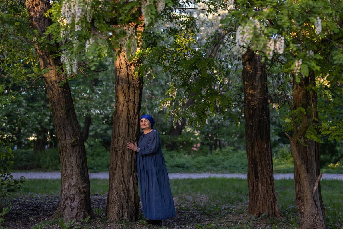 A woman in a long blue dress and head covering leans against a tree in a park