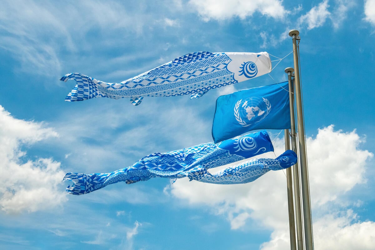 A United Nations flag on a flag pole alongside three “koinobori” (decorated carp-shaped streamers) against a blue sky with some white clouds.