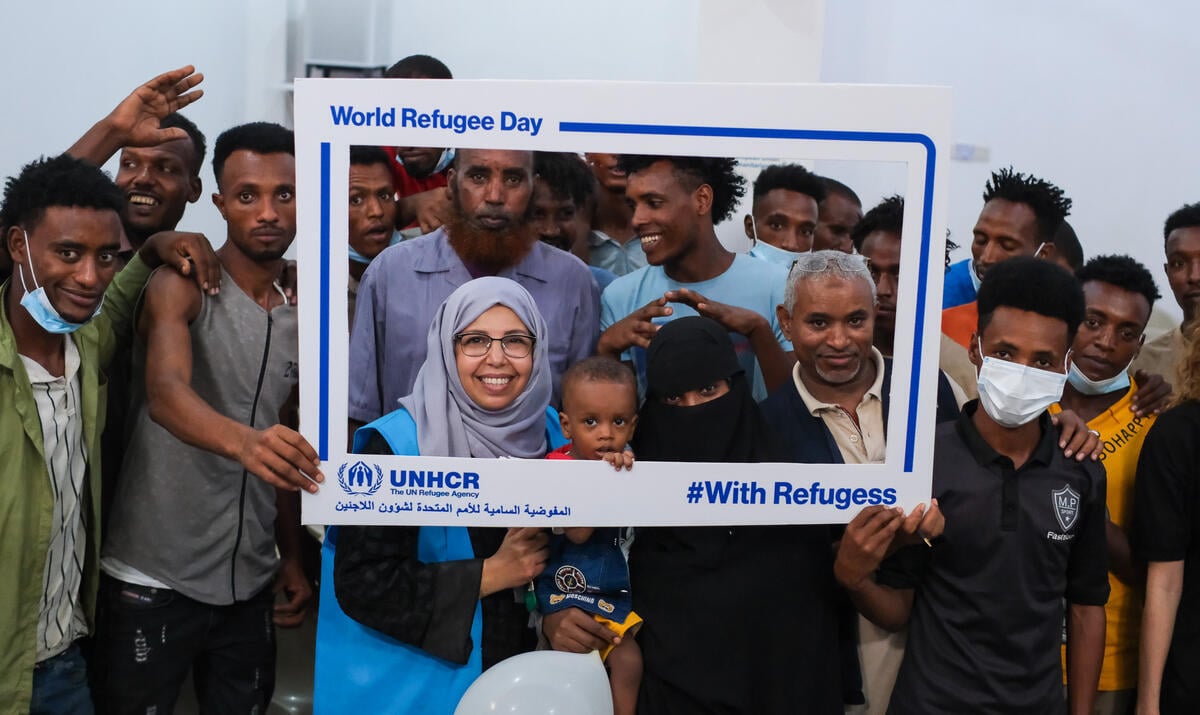 Refugees and UNHCR staff holding up a UNHCR frame which says 'World Refugee Day&quot; and '#WithRefugees&quot;