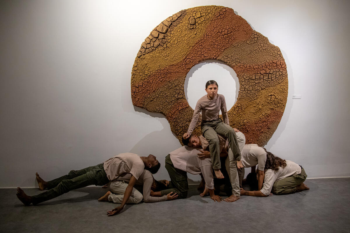 Dancers sit and lay on other crouched dancers as part of a performance. Behind them is a large work of art, a donut shape in shades of brown.