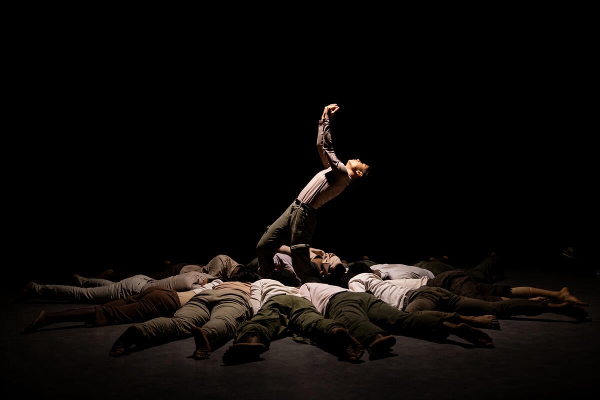 As part of a dance performance, one dancer stands, leaning back with arms in the air, in the centre of a circle of people laying face-down on the floor