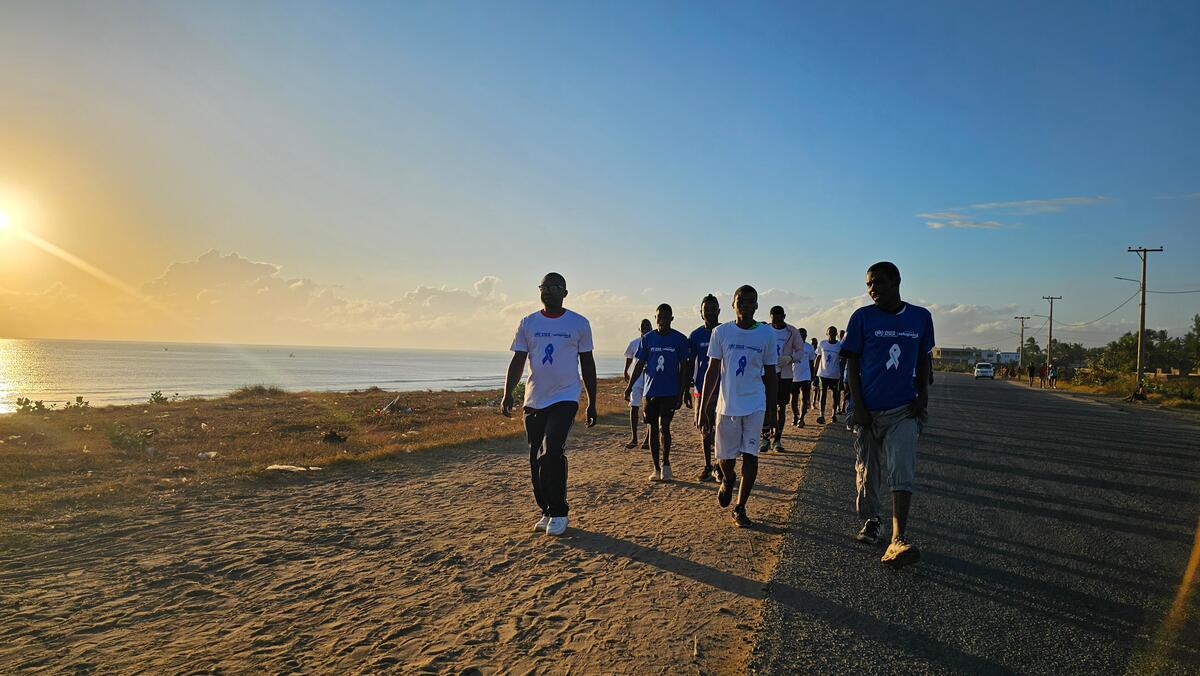 A group of people walk along a road beside a beach at sunrise.