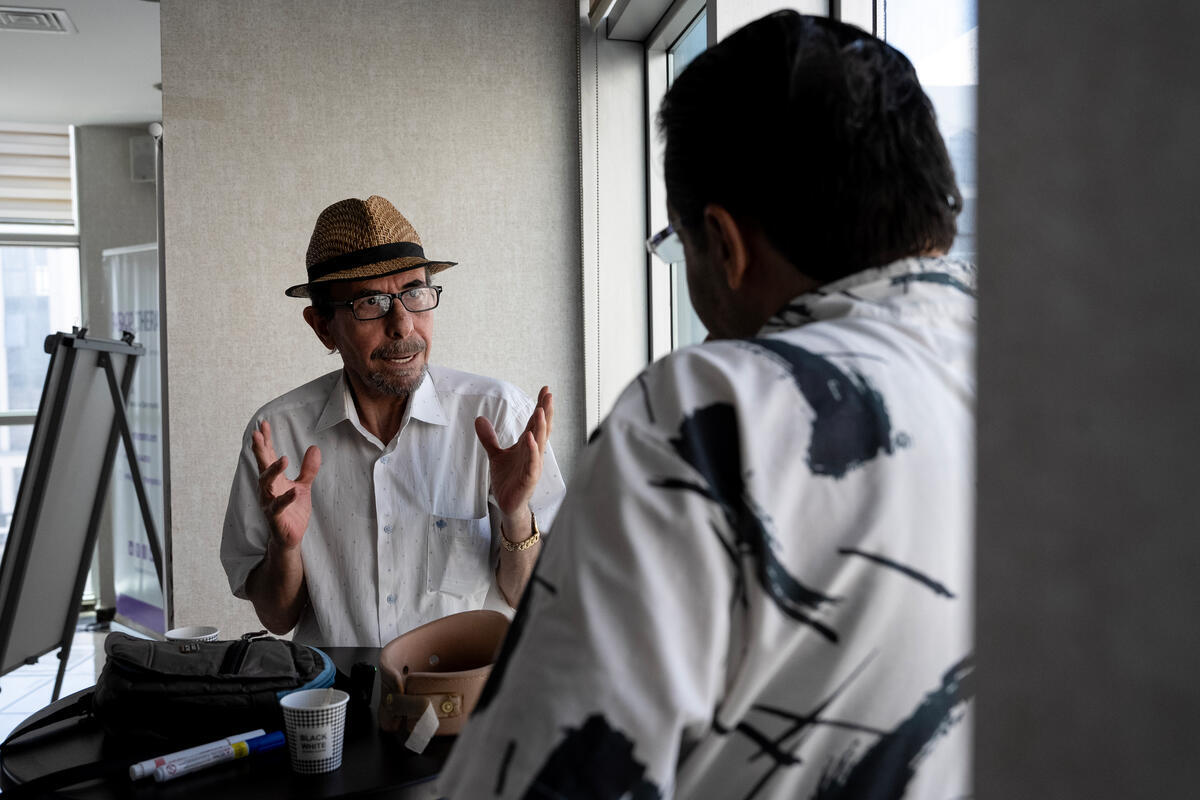 An older man in a straw trilby hat talks to another man with his back to the camera.