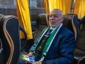 A man sits on a bus wearing a suit and scarf and holding a mobile phone