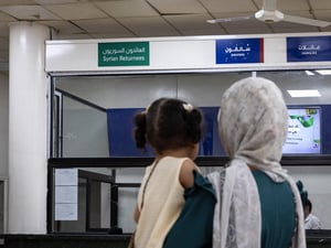 A woman carrying her daughter stands in front of a reception counter with a sign reading 'Syrian returnees' in Arabic and English
