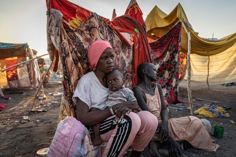 A young woman holds her baby sister with her mother sitting beside them backed by shelters made from wooden sticks and blankets.