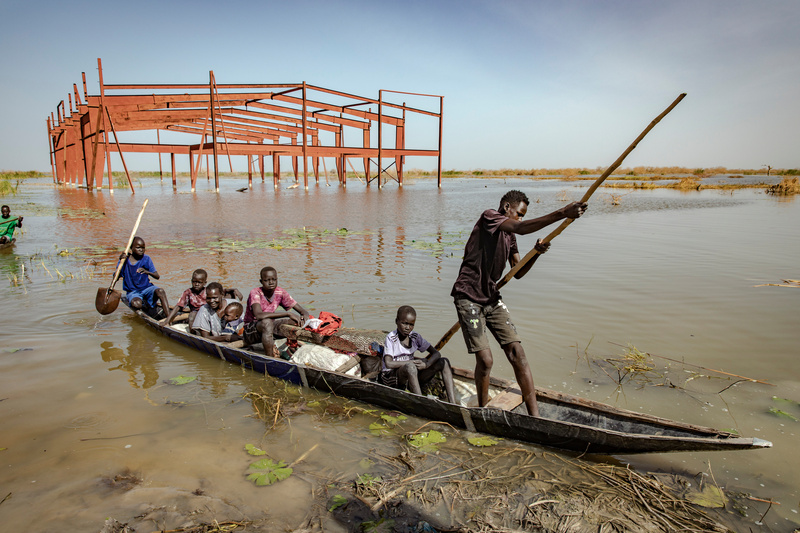 A woman and children sit in a dug out canoe being pushed through flood waters by a young man with a stick.