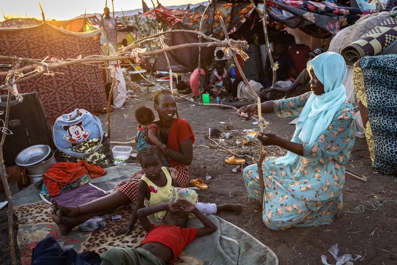 A woman seated on the ground with her three children watches another woman tie sticks together for a shelter.