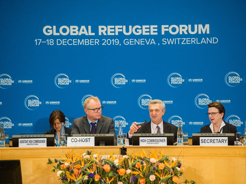 Three people (from right to left: Co-host, High Commissioner for Refugees and Secretary) sit at a podium at the Global Refugee Forum in December 2019, with one person (Assistant High Commissioner (Protection)) in the background.