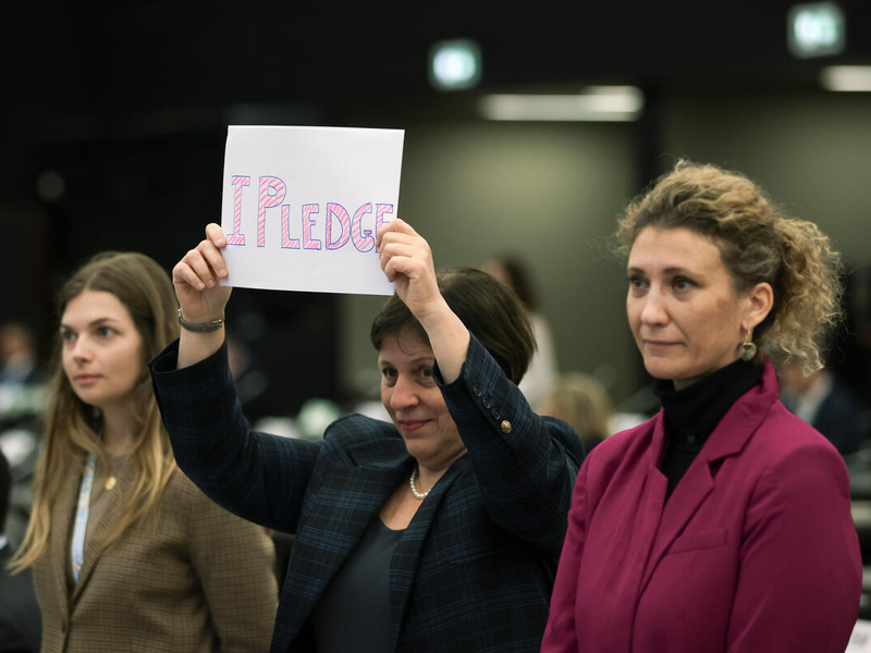 Three individuals stand together, one holds up a sign that reads 'I pledge'.