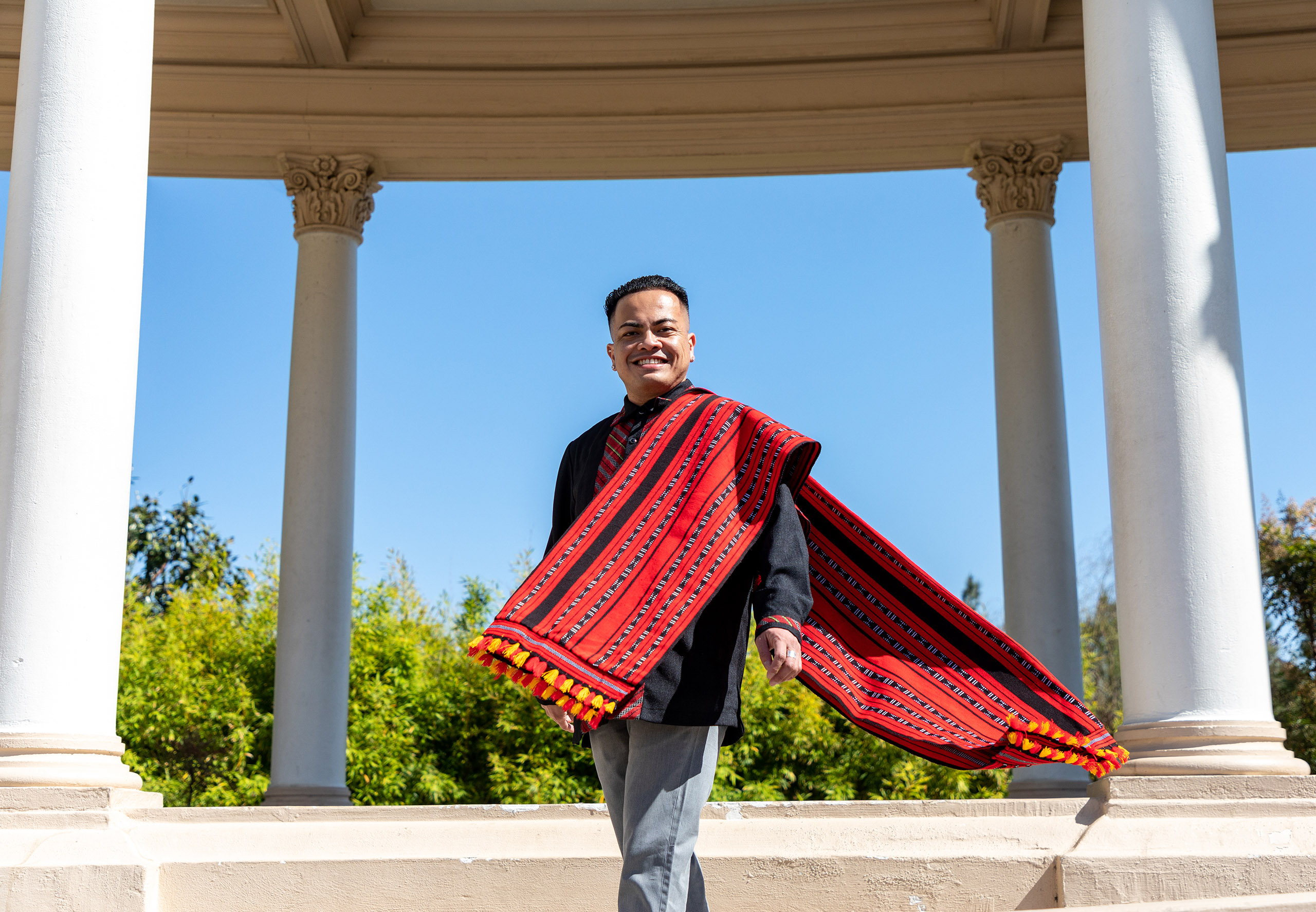 A Philippino-American man with a long, red woven scarf stands among several stone columns under a blue sky.  