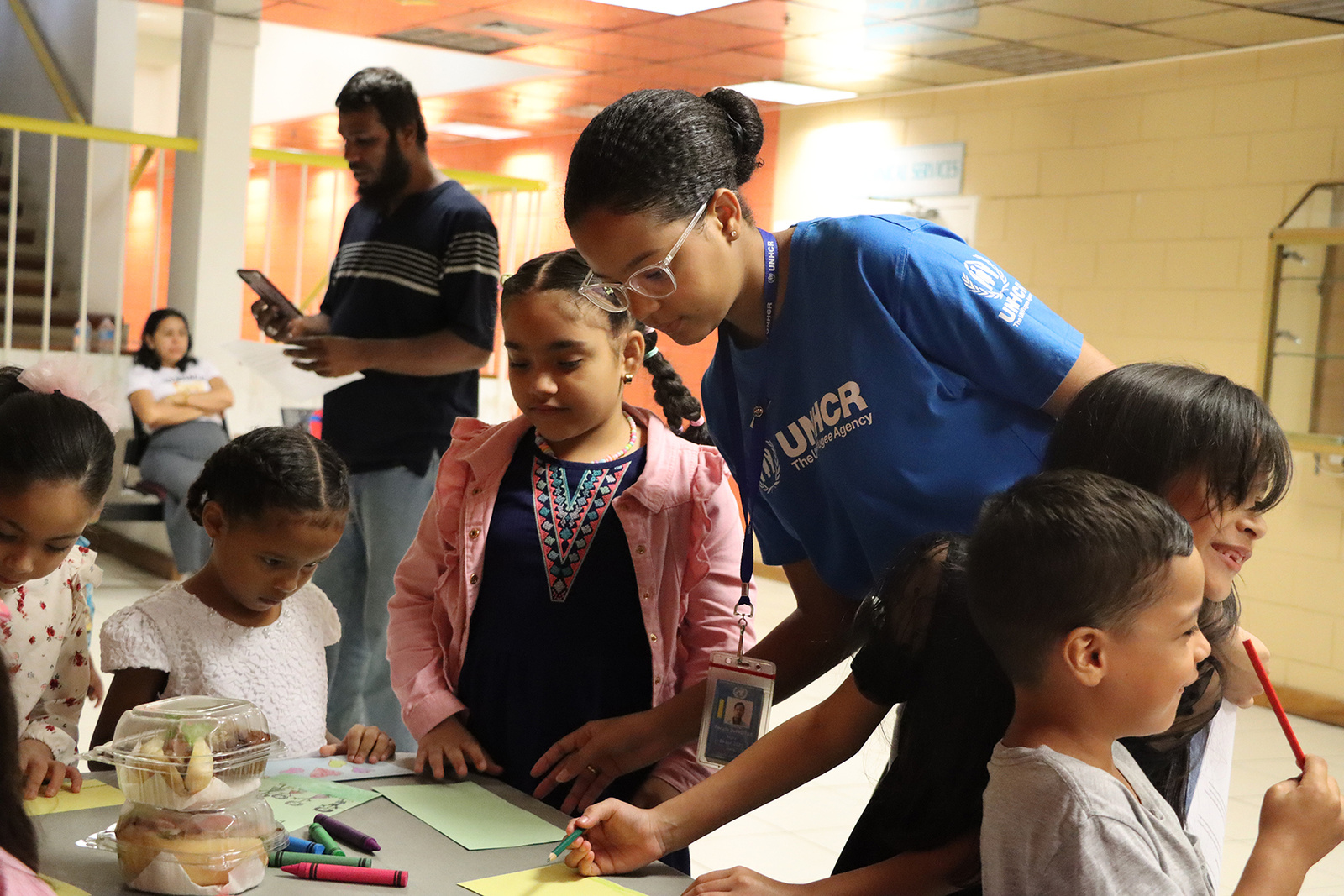 a staff member in a blue UNHCR t-shirt draws with young students around a table covered in art supplies and snacks